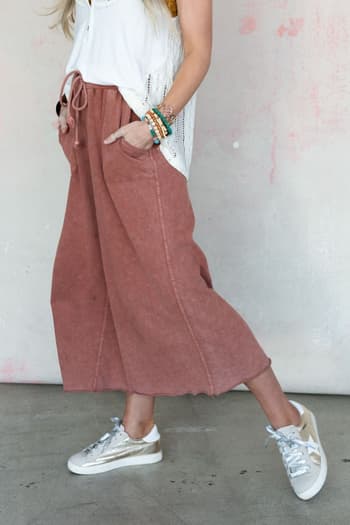 A woman with long blonde hair wears a sleeveless white top, Relaxing Robin Wide Leg Pant - Red Bean with pockets, and white sneakers. She poses indoors, showing colorful beaded bracelets on her left wrist.