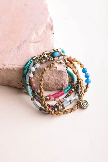 The Sunburst Stacked Bracelet Set - Multi features a lively mix of colorful beaded and braided boho bracelets with charms in blue, pink, turquoise, white, and gold, displayed on a light stone surface.