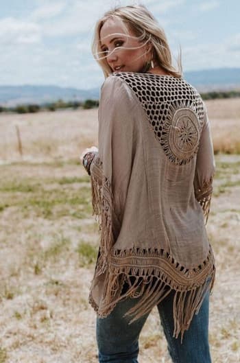 A woman stands in a grassy field, wearing the Dreamy Macramé Kimono in Mocha with bohemian crochet details and tassel fringe. She glances over her shoulder, her long blonde hair flowing under a partly cloudy sky.