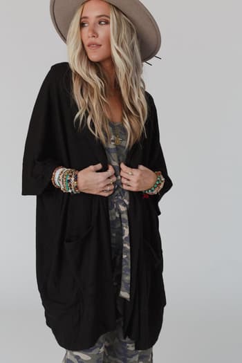 A woman with long blonde hair wears The Nest Boho Essential Hooded Ribbed Sweater in black over a camouflage outfit, styled with layered necklaces and colorful bracelets on both wrists, standing against a plain background.