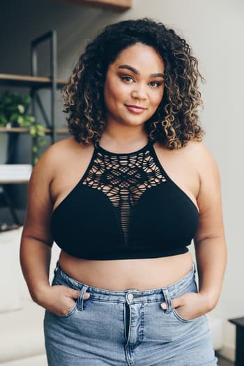 A woman with curly hair wears the Floral Cut Out High Neck Bralette in black and light blue jeans. She stands indoors, hands in her pockets and smiling softly, with a shelf of plants visible in the background.