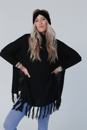 A woman with long blonde hair and a black headband models the Paloma Poncho Sweater in black, featuring an oversized silhouette with fringe, styled with blue pants and bracelets against a light gray background.