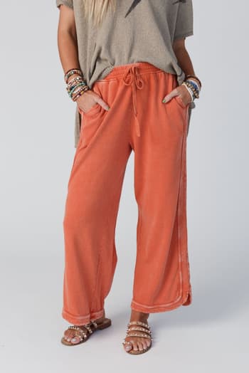 A person wearing So Comfy Wide Leg Pant Cropped Length - Faded Rust with a beige short-sleeve top, beaded bracelets, and tan studded sandals stands with hands in pockets. The face is not visible.