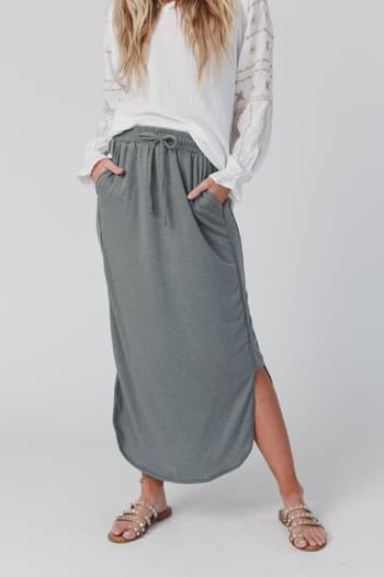 A woman stands against a white background wearing a loose white long-sleeve top, tan studded sandals, and The Nest So Comfy Drawstring Maxi Skirt in Light Olive with an elastic waistband, side slits, and her hands in the pockets.