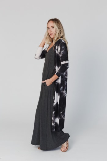 A woman with long blonde hair wears the My Go To Duster Cardigan - Black Tie Dye over a black maxi dress. She stands sideways, looking at the camera with one hand in her pocket and the other touching her hair.