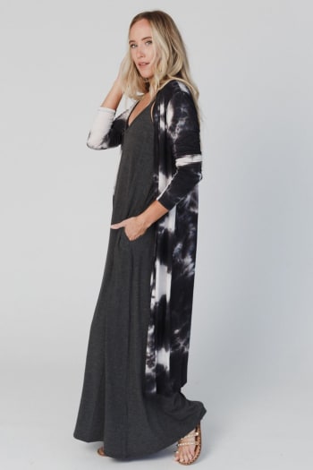 A woman with long blonde hair wears the My Go To Duster Cardigan - Black Tie Dye over a black maxi dress. She stands sideways, looking at the camera with one hand in her pocket and the other touching her hair.