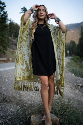 A woman stands barefoot on a tree stump by the roadside in a black dress and the Crushin' On You Velvet Kimono - Olive, featuring tassel details and bracelets. Trees and mountains are seen in the background.