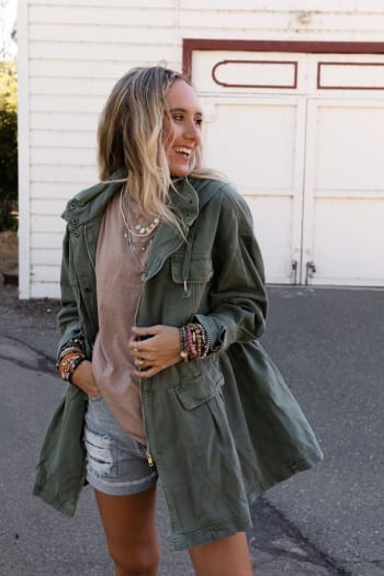 A smiling woman with long blonde hair wears the Layers On Layers Perfect Boho Parka in olive over a beige t-shirt and ripped denim shorts, accessorized with layered necklaces and bracelets, standing outside a white building.