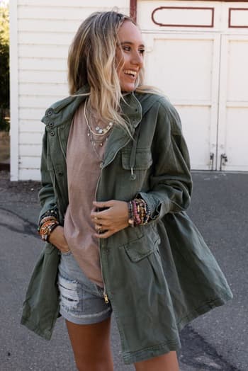 A smiling woman with long blonde hair wears the Layers On Layers Perfect Boho Parka in olive over a beige t-shirt and ripped denim shorts, accessorized with layered necklaces and bracelets, standing outside a white building.