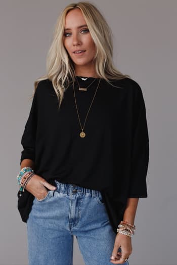 A woman with long blonde hair wears The Nest Cozy Cool Oversized Tee in black, paired with light blue jeans, layered necklaces, and colorful beaded bracelets, standing with one hand in her pocket against a neutral background.