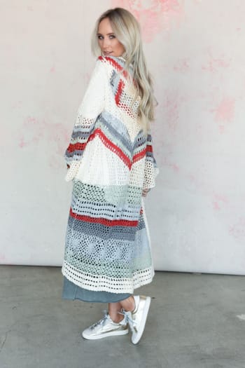 A woman with long blonde hair wears the Love In Layers Cardigan - Multi, a duster-length, loose-knit cardigan with red, gray, and white stripes over blue jeans and white sneakers, embracing bohemian style against a light wall.