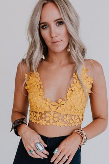 A woman with long, wavy blonde hair models the Dainty Daisy Crochet Bralette in yellow, paired with dark high-waisted pants and layered rings and bracelets, standing against a plain light background.