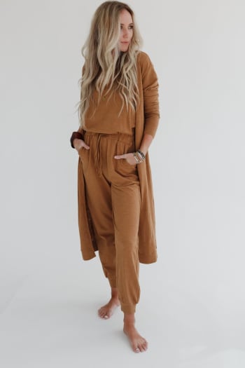 A woman with long, wavy blonde hair stands barefoot against a white background, wearing the Set to Go Textured Jumpsuit And Cardigan Set in camel, with her hands in her pockets as she looks off to the side.
