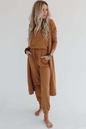 A woman with long, wavy blonde hair stands barefoot against a white background, wearing the Set to Go Textured Jumpsuit And Cardigan Set in camel, with her hands in her pockets as she looks off to the side.