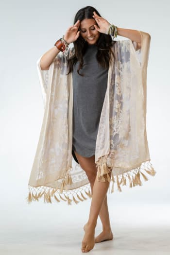 A smiling woman stands barefoot in a gray dress, layering the Crushin' On You Velvet Kimono - Nude. The sheer kimono features floral boho style and tassel details. She wears colorful bracelets, posed with her hands near her head against a white background.