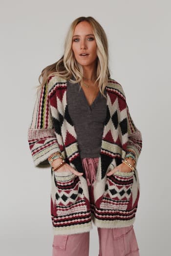 A woman with long blonde hair models the Keep It Up Oversized Cardigan - Berry, featuring red, black, and cream geometric patterns. She stands with her hands in the cardigan's pockets against a plain background.