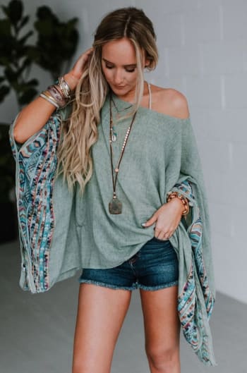 A woman wearing the Fallon Embroidered Sleeve Poncho in sage, featuring colorful sleeves, styled with layered necklaces, bracelets, and denim shorts, stands indoors looking down and touching her hair.