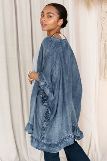 A woman with dark hair pulled back wears the Robbie Denim Jacket - Blue, an oversized fit with ruffle trim, over a grey top and jeans. She stands sideways looking over her shoulder, with neutral curtains in the background.