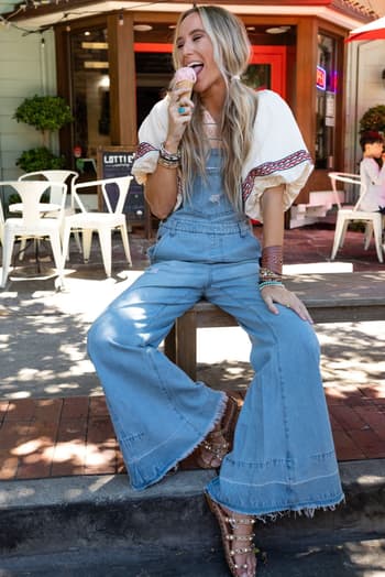 Wearing the Milly Distressed Hem Wide Leg Overall in denim and a patterned blouse, a woman sits curbside by a cafe enjoying ice cream, her boho vibe complete with layered bracelets, sandals, and loose long hair.