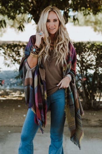 A woman with long, wavy blonde hair smiles outdoors in a Cozy Plaid Pocket Ruana Wrap - Fuchsia over her brown top and blue jeans, accessorizing with bracelets and rings amid lush bushes and trees.