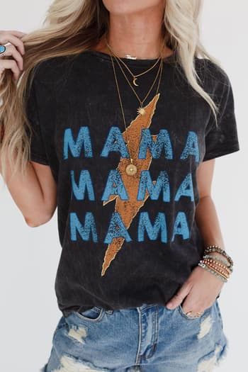 A woman wears the Thunder Mama Vintage Graphic Tee in charcoal, showcasing blue distressed "MAMA" lettering repeated three times with a gold lightning bolt. She styles it with denim shorts, layered necklaces, bracelets, and long blonde hair.