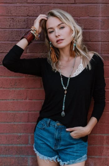 A blonde woman poses against a red brick wall wearing the Ryder Long Sleeve Bralette Tee - Black, paired with blue denim shorts, layered necklaces, large earrings, bracelets—one hand in her hair, the other in her pocket.