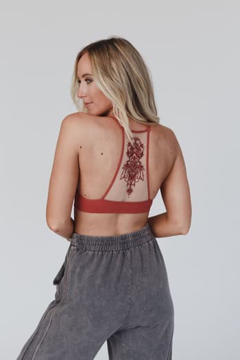 A woman with long blonde hair, facing away, wears the Tattoo Bralette - Rust and gray sweatpants, showcasing a large floral tattoo on her upper back against a plain light background.