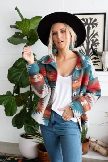 A woman in a wide-brimmed black hat, Anika Mixed Pattern Jacket - Teal, white top, and jeans stands indoors by potted plants and wall art, gazing at the camera with a relaxed expression.