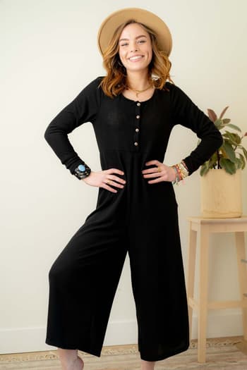 A smiling woman radiates effortless style indoors, standing hands-on-hips by a table with a potted plant. She wears the Simply Comfort Jumpsuit - Black, a scoop-neck boho piece, paired with colorful bracelets and a wide-brimmed hat.