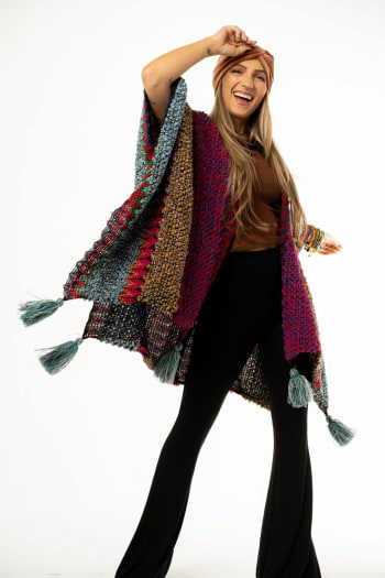 A smiling woman poses against a white background, wearing the Desert Dreams Crochet Ruana - Magenta with vibrant knit fabric and tassels. She pairs it with a brown top, black pants, headband, bracelets and lifts one arm in playful boho style.