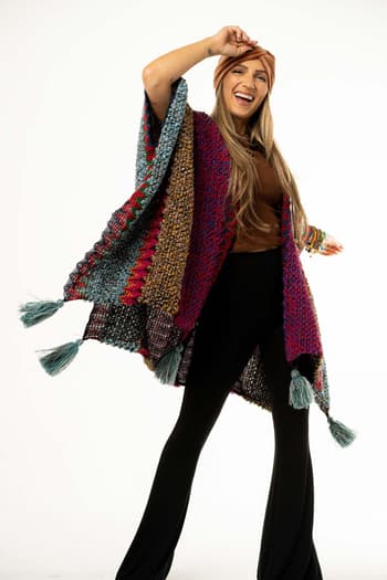 A smiling woman poses against a white background, wearing the Desert Dreams Crochet Ruana - Magenta with vibrant knit fabric and tassels. She pairs it with a brown top, black pants, headband, bracelets and lifts one arm in playful boho style.