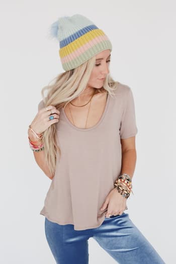 A woman in a relaxed fit Scoop Neck Bralette Tee - Khaki, blue velvet pants, a pastel striped knit beanie, and colorful bracelets looks down while touching her hair against a plain white background.