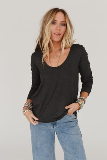 A woman with long, wavy blonde hair wears the Ryder Long Sleeve Bralette Tee in Charcoal and light blue jeans, accessorized with bracelets and a necklace, standing against a plain light background.
