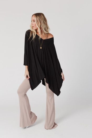A woman with long blonde hair wears The Nest The Wren Top in black, styled with beige flared pants and a gold necklace, standing against a plain white background.