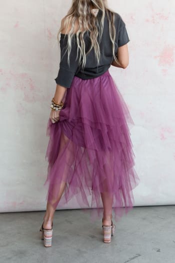 A person with long, wavy hair poses by a light wall in a dark top and the Boho Ballerina Skirt - Mauve. Lifting the layered tulle skirt, they show high-heeled sandals, creating a casual and playful look.