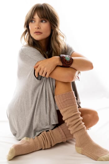 A woman with long, wavy hair sits on the floor in a relaxed pose, wearing Cozy Cabin Lounge Socks - Pink and a loose gray t-shirt. She gazes thoughtfully to the side against a plain white background.
