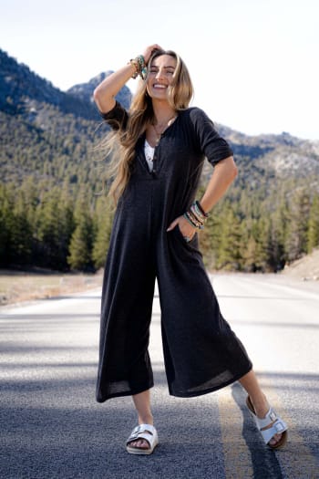 A smiling woman stands on a sunny mountain road in the Simply Comfort Jumpsuit - Dark Gray, a scoop neck boho style with an oversize silhouette. She pairs it with white sandals and colorful bracelets, framed by pine trees and rolling hills.