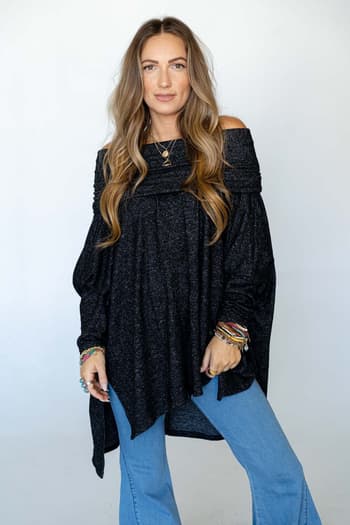 A woman with long wavy hair wears The Nest So Versatile Flowy Knit Sweater in charcoal, styled boho with light blue jeans, layered necklaces, and colorful bracelets, standing before a plain white background.
