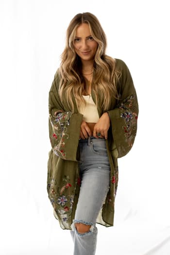 A woman with long, wavy blonde hair models the Fun In Florals Kimono - Olive over a white top and ripped light blue jeans, standing in front of a white background and smiling slightly.