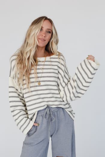 A woman with long blonde hair stands against a white background, smiling slightly with her left hand in her pocket, wearing the All My Stripes Oversized Sweater - Ivory Gray and loose gray pants.