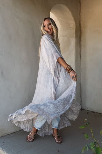 A woman with long blonde hair smiles while twirling in the Paisley Tapestry Free Flow Duster Kimono - Gray, styled over jeans and sandals. She stands by a textured wall with an arched recess, sunlight casting soft shadows.