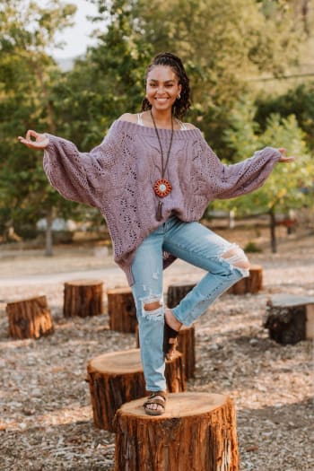 A smiling woman wears the Golden Hour Bubble Sleeve Sweater Dress in Lavender, standing on one foot atop a tree stump with arms outstretched among trees and more stumps in the background.