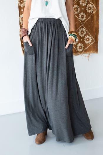 Indoors, someone wears a white top and The Perfect Pocketed Maxi Skirt - Charcoal, with real pockets and a stretch waist. They style it with bracelets, a turquoise necklace, brown ankle boots, and a patterned brown wall hanging behind them.
