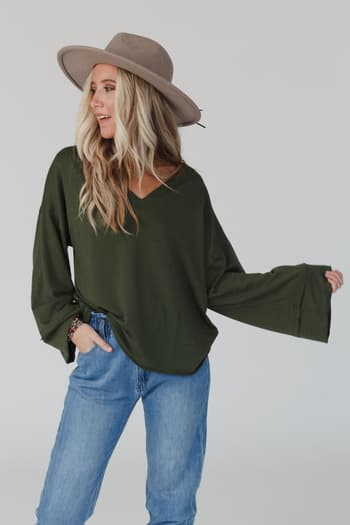 A woman wears The Nest Love It V Neckline Sweater Top in olive, a wide-brimmed hat, and light blue jeans, posing with one hand in her pocket against a plain light background.