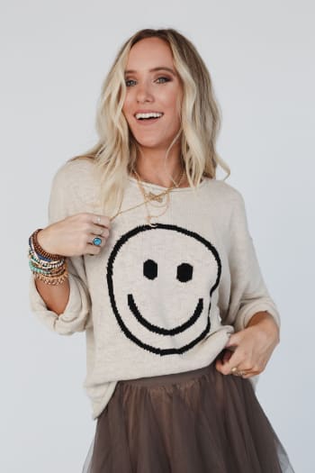 A smiling woman with long blonde hair wears the Made You Smile Sweater - Ivory, layered necklaces, beaded bracelets, and a brown tulle skirt, posing against a plain light background.
