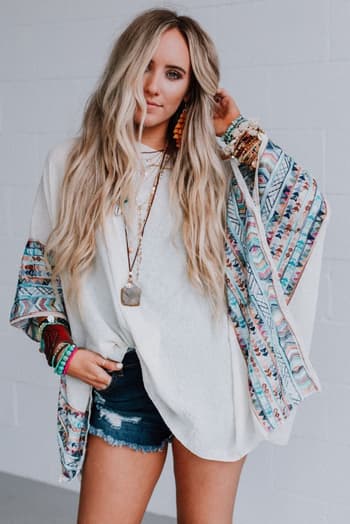 A woman with long, wavy blonde hair wears the Fallon Embroidered Sleeve Poncho in ivory, paired with denim shorts, layered necklaces, and multiple bracelets, standing against a light gray brick wall.