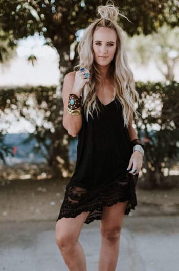 A woman with long, wavy blonde hair wears the Prima Lace Hem Slip in black, accessorized with bracelets and rings, posing confidently outdoors against a backdrop of leafy trees.