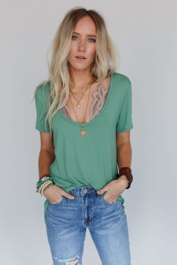 A woman with long blonde hair wears the Scoop Neck Bralette Tee in Sage, styled with layered necklaces, bracelets, and blue jeans. She stands against a plain light gray background with her hands in her pockets.