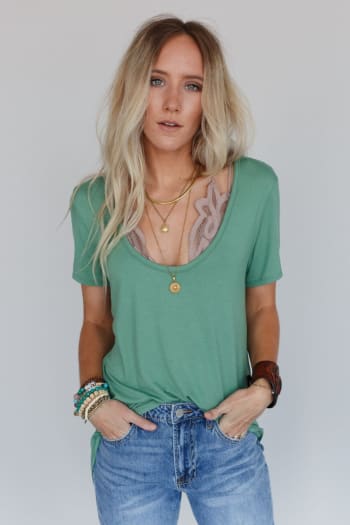 A woman with long blonde hair wears the Scoop Neck Bralette Tee in Sage, styled with layered necklaces, bracelets, and blue jeans. She stands against a plain light gray background with her hands in her pockets.