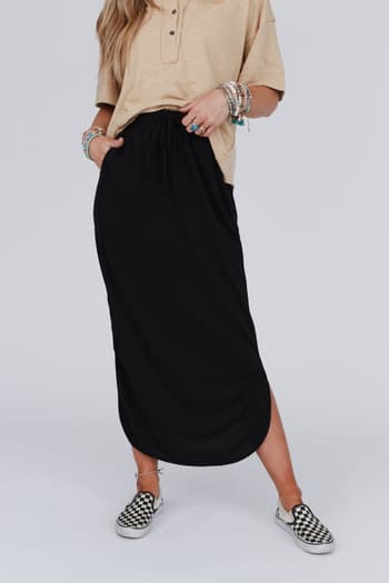 A person wears a beige short-sleeve shirt, multiple bracelets, black-and-white checkered slip-on shoes, and The Nest So Comfy Drawstring Maxi Skirt in black while standing against a plain light background.